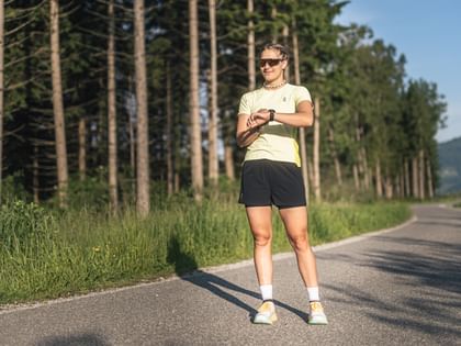 Female runner stands on a paved path at the edge of a forest, looking at her sports watch in sunny conditions.
