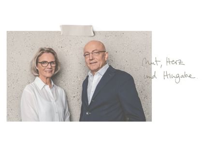 Eine Frau mit Brille und ein Mann im Anzug stehen lächelnd nebeneinander vor einer hellen Wand. Daneben steht handschriftlich „Mut, Herz und Hingabe.“