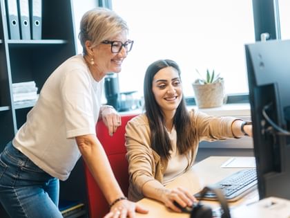 One woman is sitting at a computer pointing at the screen, while another woman stands beside her smiling. On the desk are a monitor, keyboard, and mouse, and in the background there are folders and a plant.