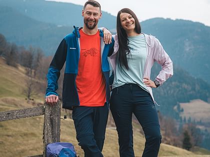 A man and a woman are standing smiling on a grassy field next to a wooden fence in a mountain landscape. A blue backpack stands in front of them, with hills and a cloudy sky visible in the background.