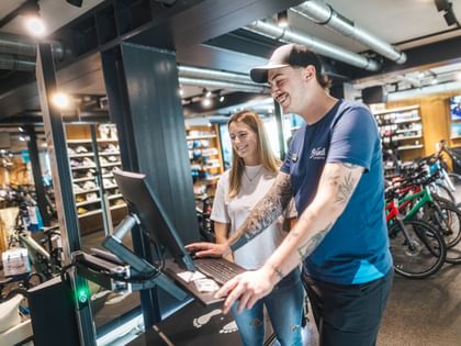 A Bründl Sports employee is carrying out a bike fitting using a digital station while a customer stands beside him and smiles. Bicycles, e-bikes and sports accessories are visible in the shop background; the Bründl Sports logo can be seen on the shirt and a bag.
