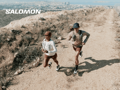Two female runners are running on a dry trail overlooking a city. The white Salomon logo is visible in the upper left corner.