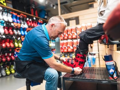 Ein Bründl Sports Mitarbeiter passt einer Kundin im Shop einen roten Skischuh an. Er sitzt vor einer Wand mit Skischuhen und sorgt für die perfekte Passform und optimalen Komfort.