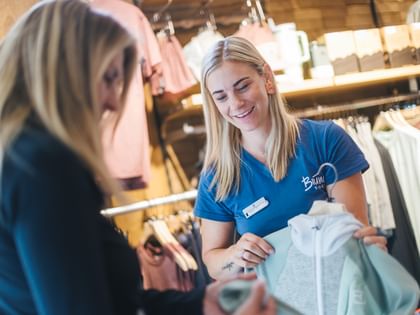 Eine Bründl Sports Mitarbeiterin zeigt einer Kundin eine helle Sportjacke auf einem Kleiderbügel. Im Hintergrund sind Sportbekleidung, Kleiderstangen und Regale mit Accessoires zu sehen.
