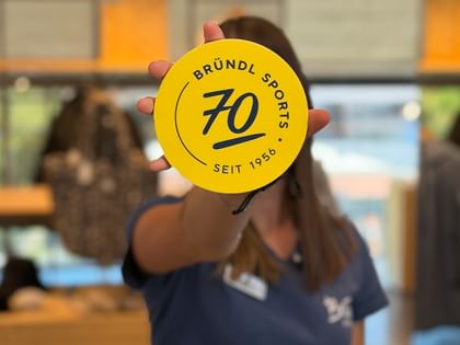 A hand holds a round yellow token displaying “Bründl Sports”, “70” and “Seit 1956” (since 1956). Ski poles and the illuminated interior of a sports shop are visible in the blurred background.