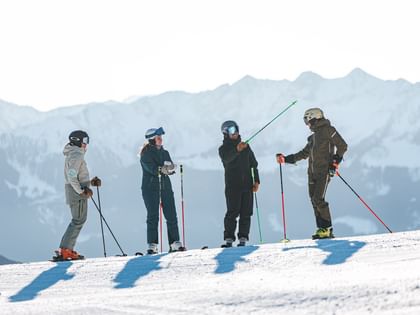 Vier Skifahrer stehen mit Ski und Skistöcken auf einer verschneiten Piste und unterhalten sich. Im Hintergrund sind schneebedeckte Berge zu sehen.