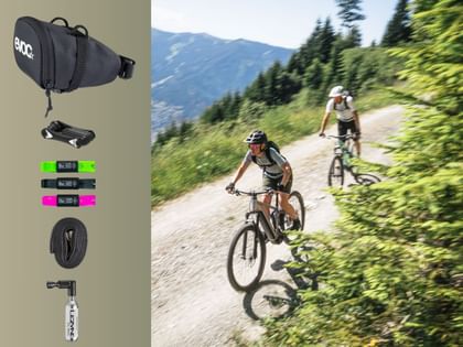 Links sind verschiedene Fahrradzubehörteile vor einem hellen Hintergrund angeordnet, darunter eine schwarze Satteltasche mit dem Schriftzug „evoc“, ein Multitool, drei Reifenheber mit der Aufschrift „Muc-Off“, ein Fahrradschlauch und eine CO₂-Kartusche mit Ventilaufsatz. Rechts fahren zwei Personen mit Helmen auf Mountainbikes einen Schotterweg durch eine grüne Berglandschaft mit Bäumen und Bergen im Hintergrund.