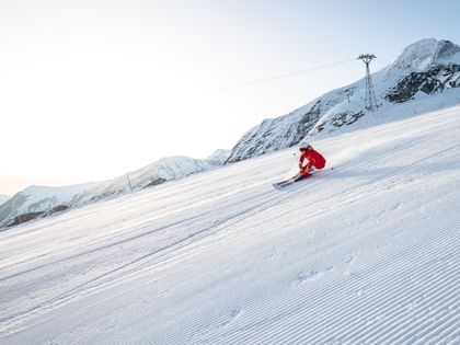 Das Bild zeigt eine Skifahrerin in roter Kleidung, die mit hoher Geschwindigkeit eine frisch präparierte Piste hinunterfährt. Der Schnee wirbelt hinter ihr auf, und im Hintergrund sind hohe Berge zu sehen.