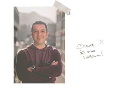 Ein lächelnder Mann im Kapuzenpullover steht mit verschränkten Armen vor einer unscharfen Bergkulisse. Daneben steht handschriftlich „DANKE für euer Vertrauen!“.