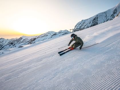 Das Bild zeigt einen Skifahrer, der bei Sonnenaufgang eine schneebedeckte Piste hinunterfährt, umgeben von Berglandschaft.