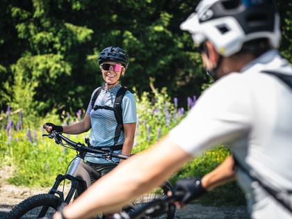 Eine Person mit Helm, Sonnenbrille und Rucksack sitzt auf einem Mountainbike und lächelt zu einer anderen Person, die im Vordergrund unscharf zu sehen ist. Im Hintergrund stehen dichte grüne Bäume und blühende Pflanzen entlang eines Waldweges.