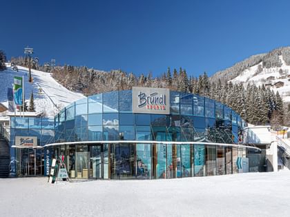 The image shows the Bründl Sports AreitXpress valley station shop with a large blue glass facade next to a snowy ski slope. The “Bründl Sports” logo and the texts “SKIDEPOT,” “SHOP,” “RENT,” “DEPOT,” and “SERVICE” are visible on the building, with a ski lift, forest, and snow-covered mountains in the background.