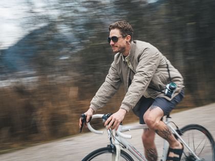 A man rides a bicycle along a path, wearing a camera around his neck and sunglasses. The background is motion-blurred and shows trees and a natural landscape.