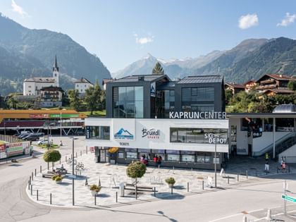 Das Kaprun Center des Kitzsteinhorns vor einer alpinen Bergkulisse. Im Gebäude befindet sich ein Bründl Sports Shop; sichtbar sind unter anderem die Schriftzüge „KAPRUN CENTER“, „Bründl Sports“, „Kitzsteinhorn“, „Tickets“ und „Ski Depot“.