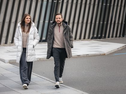 Zwei Personen in nachhaltiger Wintermode von Ecoalf spazieren entspannt durch die Stadt – moderne Daunenmäntel in Weiß und Grau für Damen und Herren.