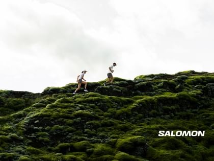 Das Bild zeigt zwei Trailrunner, die über eine mit leuchtend grünem Moos bewachsene Felslandschaft laufen, während der Himmel über ihnen hell und wolkig erscheint; das „Salomon“-Logo ist dezent in der unteren rechten Ecke platziert.