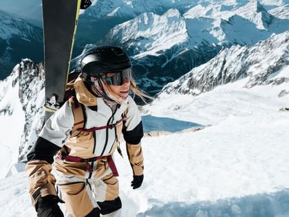 Peak Performance AW 25 A skier climbs a snowy mountain slope with skis strapped to her backpack. She wears a beige and black ski jacket, helmet, and goggles, with majestic mountain peaks in the background.