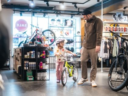 Das Bild zeigt ein kleines Mädchen in der Bründl Sports Bikeworld, das ein Kinderfahrrad schiebt und dabei fröhlich mit einem Erwachsenen spricht.
