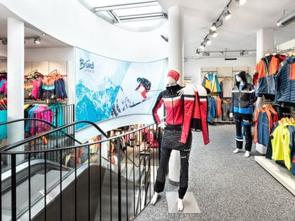 Bründl Sports Saalbach Life.Style - Innenansicht - Rechts im Bild Skitourenoutfits, in der Mitte eine Rolltreppe ins untere Stockwerk und Ski Textil für Kinder links