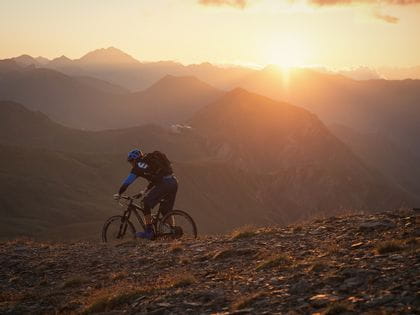 ein Mountainbiker fährt durch die Berglandschaft, während im Hintegrund die Sonne untergeht