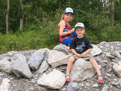 Zwei Kinder sitzen lächelnd auf großen Felsen, beide tragen Bründl Sports-Kappen. Sie sind in einer natürlichen Umgebung mit grünen Bäumen im Hintergrund, was die Freude an Outdoor-Aktivitäten und die Schönheit der Natur unterstreicht.