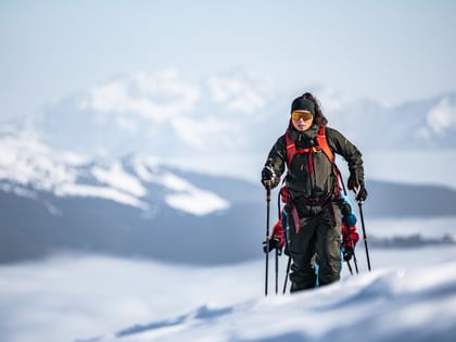 A person in ski touring gear ascends a snowy slope, with an impressive mountain landscape and cloud-covered valleys visible in the background.