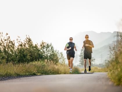 Two people jogging side by side on a tarmac path, surrounded by meadows and trees. In the background, hills and electricity pylons can be seen in the light of the morning sun.