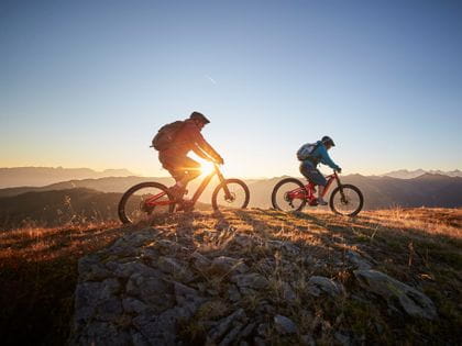 zwei Mountainbiker fahren auf einem Berggrat, im Hintergrund der Sonnenuntergang