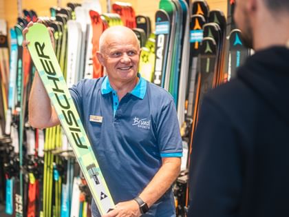 Manfred Rogetzer steht in Bründl Sports Kleidung vor einer Wand mit Skiern, hält ein Paar Fischer-Ski in der Hand und spricht mit einer Person im Vordergrund.