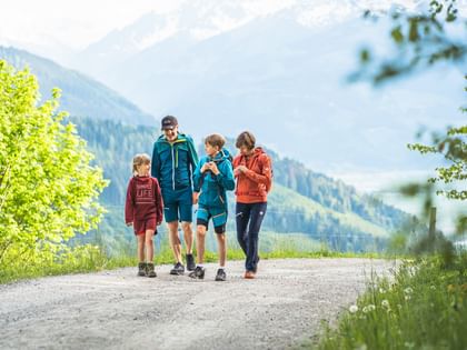 Das Bild zeigt eine Familie beim Wandern auf einem idyllischen Bergweg. Die beiden Erwachsenen und zwei Kinder tragen Outdoor-Kleidung und spazieren entspannt auf einem Kiesweg, umgeben von grünen Bäumen und Hügeln im Hintergrund. Sie wirken glücklich und entspannt, was die Freude an der Natur und dem gemeinsamen Abenteuer unterstreicht.