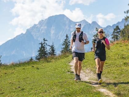 A woman and a man are running side by side on a narrow path through a green meadow. In the background, tall mountains, a few trees, and a sky with white clouds are visible.