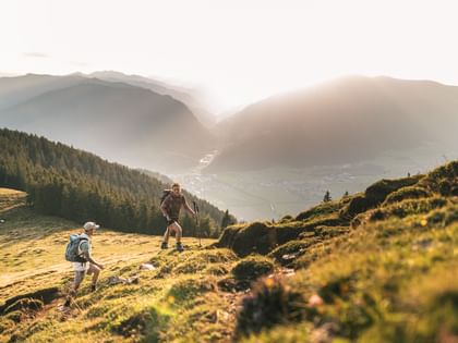Zwei Personen mit Rucksäcken steigen einen steilen, grasbewachsenen Hang hinauf. Im Hintergrund liegt ein Tal mit einem kleinen Ort, umgeben von bewaldeten Hügeln und Bergen im warmen Licht des Sonnenuntergangs.