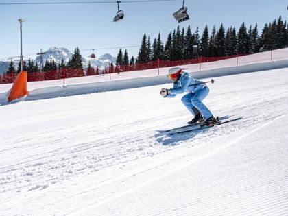 A person in light blue ski clothing skis in a crouched position down a snowy slope. A red safety net, gondolas, fir trees and snow-covered mountains are visible in the background.