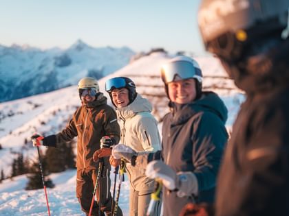 Vier Skifahrer stehen mit Helm, Skibrille und Skistöcken zusammen auf einer verschneiten Piste und lächeln. Im Hintergrund sind schneebedeckte Berge im warmen Morgenlicht zu sehen.