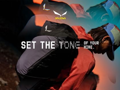 A person wearing a cap and red clothing carries a black backpack and leans forward. The image includes the text “SET THE TONE OF YOUR HIKE.” and the brand logo “SALEWA.”