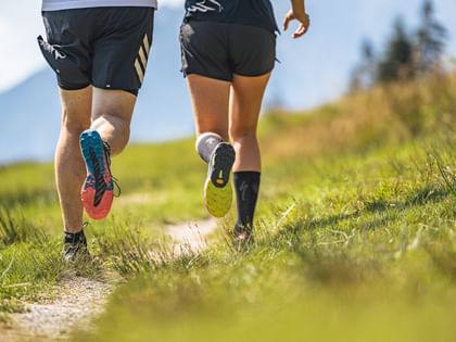 Die Beine von zwei laufenden Personen sind von hinten zu sehen, während sie auf einem schmalen Pfad durch grünes Gras laufen. Die Sohlen der Laufschuhe sind sichtbar, und die Umgebung besteht aus einer sonnigen Wiese.
