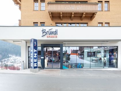 Das Bild zeigt den Eingang eines Bründl Sports Shops in einem alpinen Gebäude. Über dem Eingang befindet sich das Logo, und der Schriftzug "RENT" weist auf die Verleihmöglichkeit hin.