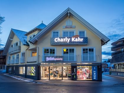 Das Bild zeigt ein Gebäude mit dem "Bründl Sports" Shop im Erdgeschoss und einem Schild für "Charly Kahr" im oberen Bereich, umgeben von anderen Gebäuden in einer winterlichen Umgebung.