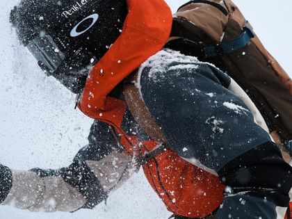 Peak Performance AW 25 A skier wearing a red and black jacket, helmet, and goggles rides through fresh powder snow. Snow sprays in all directions as they move dynamically.