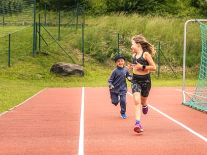 In the picture, two children are running on a track - a girl in front in sportswear, a smaller boy with a cap and tracksuit behind her. Both look at each other with a smile and move towards a football goal at the edge of the track.