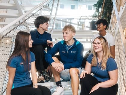 Five young people sit on a staircase inside a building, looking at each other while talking. Some of them wear blue shirts with the text “Bründl Sports.”
