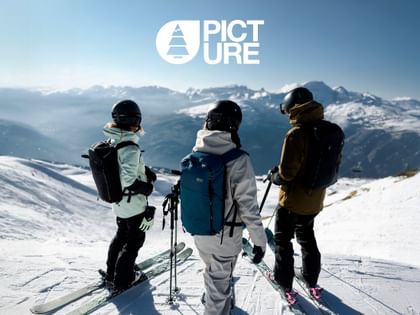Drei Skifahrer:innen in moderner Ski- und Outdoor-Bekleidung von Picture stehen auf einem verschneiten Gipfel und blicken über die winterliche Berglandschaft.