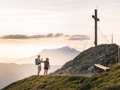 A woman and a man with backpacks stand on a mountain summit and give each other a high-five next to a large wooden cross. In the background are mountains, a sky with clouds, and warm evening light.