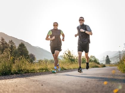 Two runners jog side by side on a paved path through an alpine landscape with low sunlight.