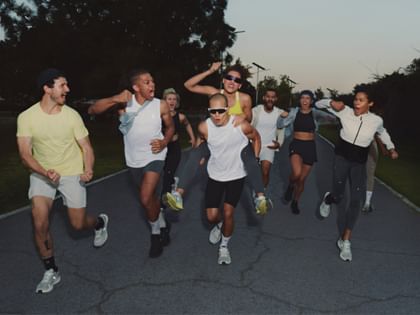A group of athletically dressed people is running with energy and joy on a road at dusk, some cheering and laughing.