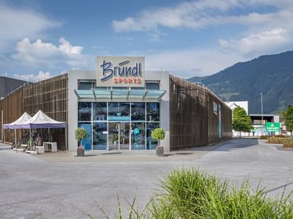 Außenansicht der Bründl Sports Filiale in Saalfelden. Vor dem modernen Geschäftsgebäude mit großem Bründl Sports Logo stehen mehrere Fahrräder unter zwei weißen Zelten. Im Hintergrund sind Berge, Bäume und ein teilweise bewölkter Himmel zu sehen. Sichtbarer Text: „Bründl Sports“ und „Einfahrt“.