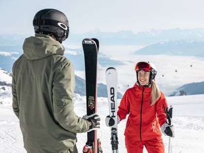 Eine Frau in roter Skiausrüstung und ein Mann in grüner Skiausrüstung unterhalten sich auf der Piste, beide halten ihre Skier.