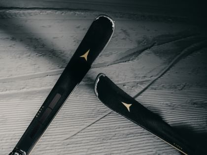 Nahaufnahme von zwei schwarzen Atomic Cloud Ski mit goldfarbenen Logos, die diagonal über einer präparierten Skipiste liegen. Die gerillte Schneeoberfläche und die Skispuren sind deutlich sichtbar.
