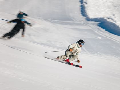 Drei Skifahrer fahren mit sportlichen Schwüngen eine verschneite Skipiste hinab. Der vordere Skifahrer trägt helle Skibekleidung und fährt auf roten Ski.