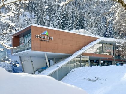 Das Bild zeigt das Bründl Sports Geschäft bei der Talstation der Schmittenbahn in Zell am See, umgeben von verschneiten Bäumen und winterlicher Landschaft. Das Gebäude ist modern und klar gestaltet.